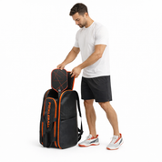 Male pickleball player in athletic wear sliding a paddle into the main compartment of a large black and orange PickleballDoor pickleball backpack against a white studio background